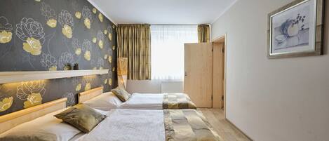 A hotel room with two beds, a large window with curtains, a wooden wardrobe, and a framed picture on the wall.