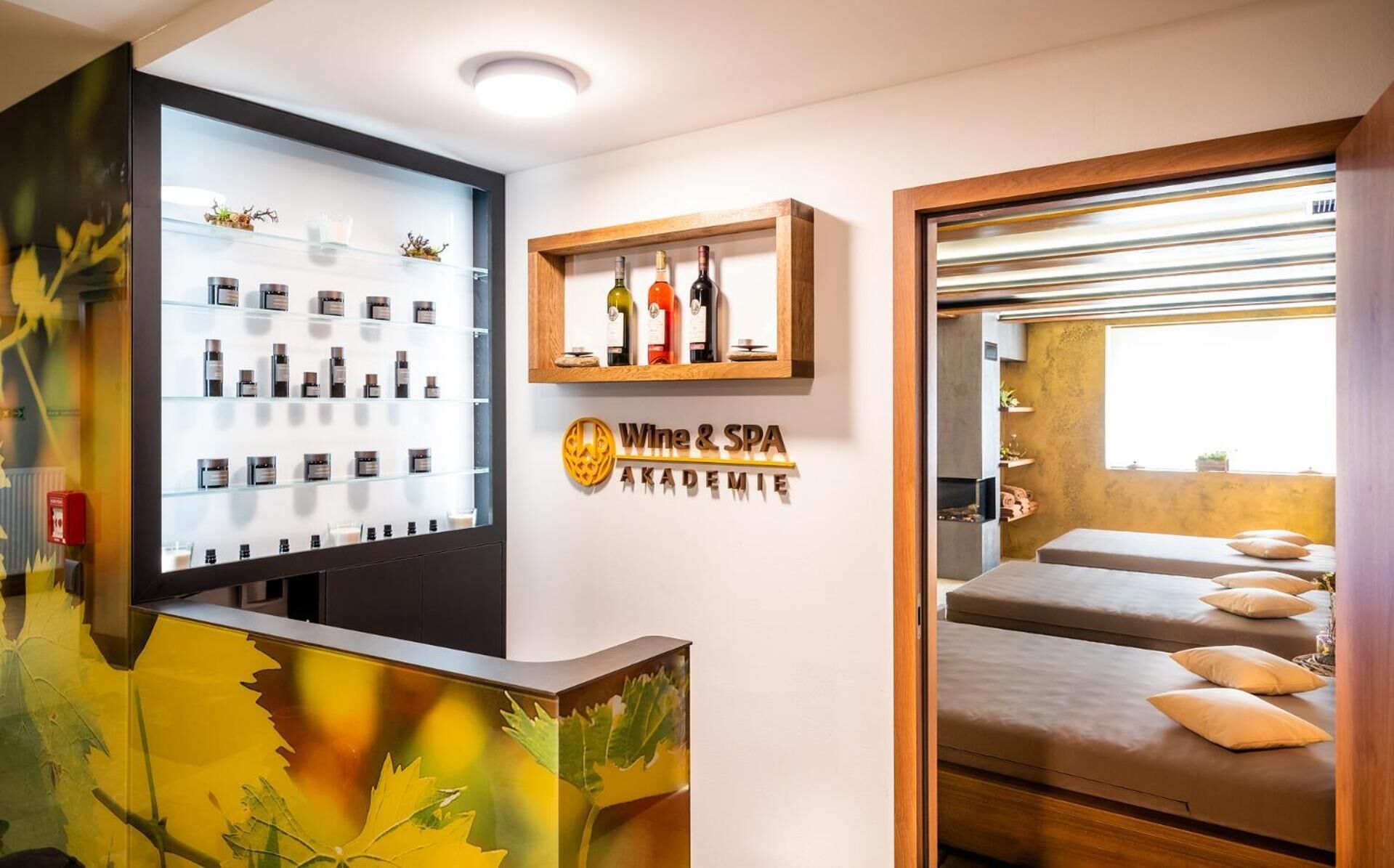 A spa area with a reception desk displaying various products, a wall-mounted sign for 'Wine & SPA ACADEMIE', and a relaxation area with beds and pillows.