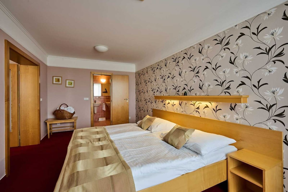 Habitación de hotel con una cama grande, cabecero de madera, una mesita de noche, alfombra roja, un banco de madera, pared con papel tapiz floral, y un baño visible a través de una puerta abierta.
