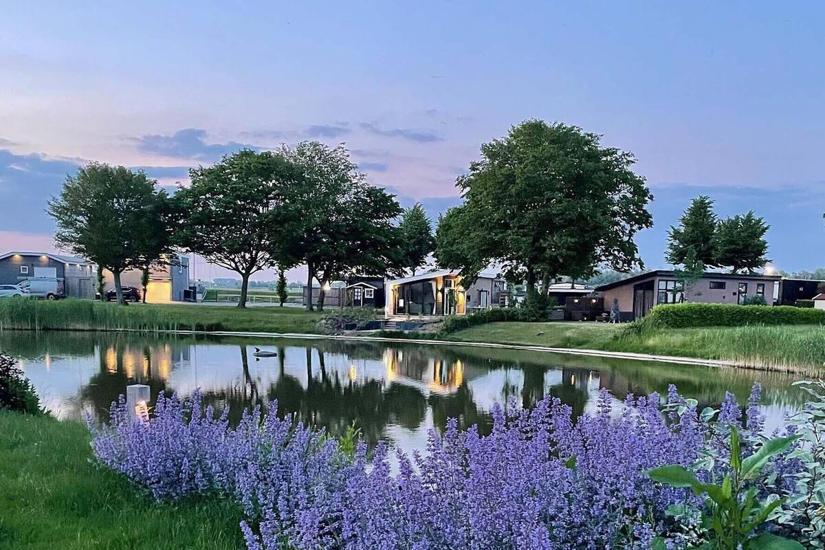 Eine ruhige Seelage mit modernen Häusern, einem stillen Teich und blühenden violetten Blumen im Vordergrund.