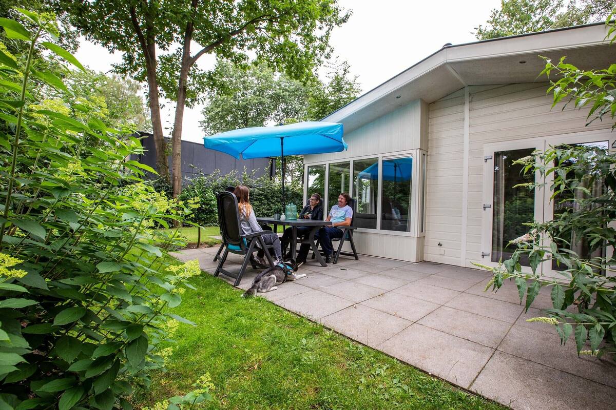Eine Terrasse im Freien mit Tisch und Stühlen, umgeben von Grünpflanzen und einem Gebäude mit großen Fenstern.