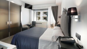 A hotel room with a bed, a desk with a telephone, a chair, a small table with a lamp, and a window with curtains. - Barbarella Suite (Naples)