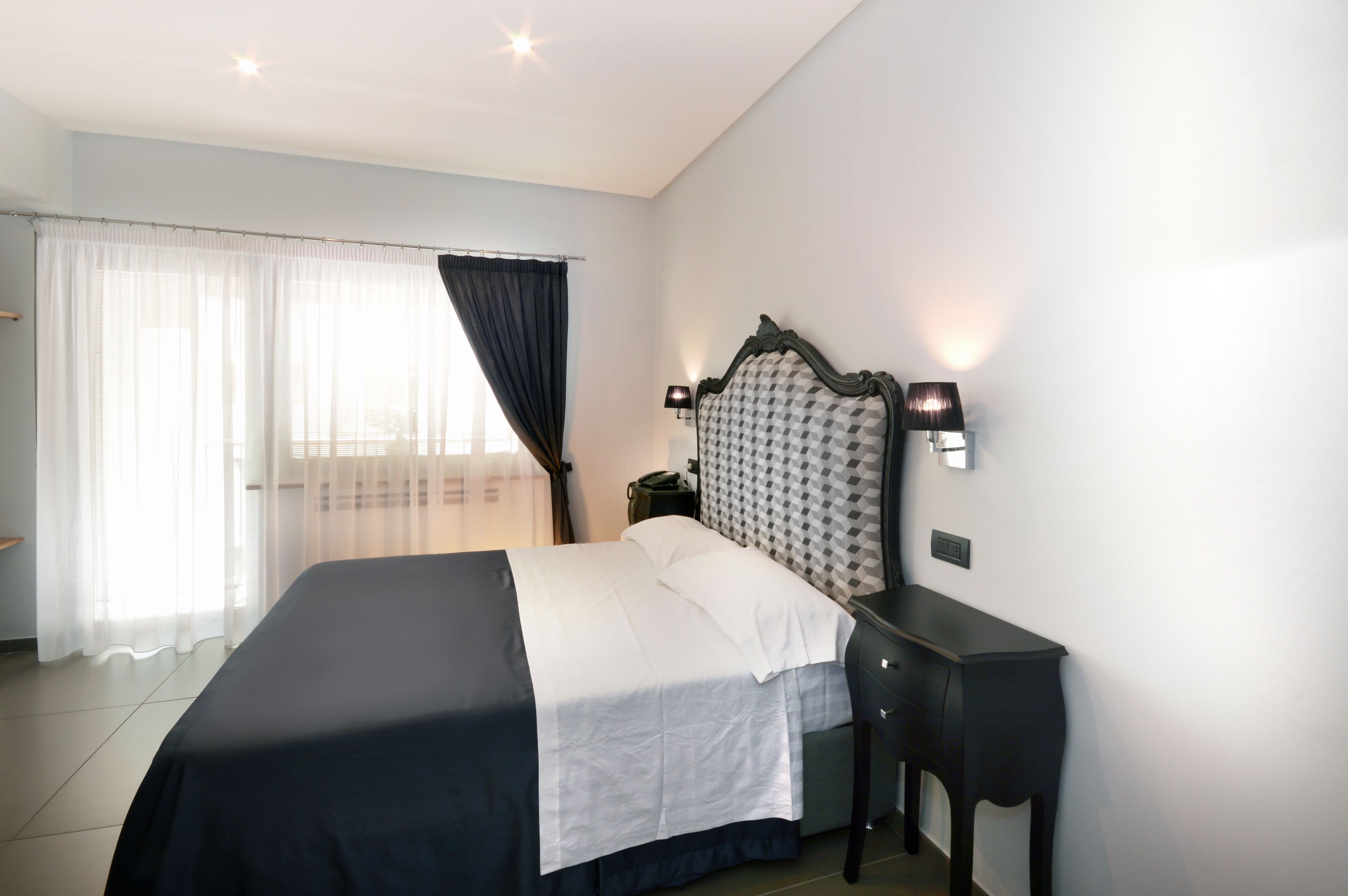 A hotel room with a bed, a nightstand, a lamp, and a window with sheer curtains.