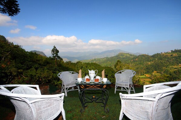A table set with white wicker chairs, a white pitcher, cups, and a teapot, overlooking a mountainous landscape. - The Hideaway Ella (Makulella)