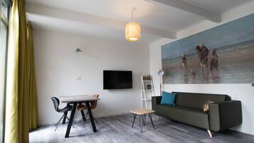 Una sala de estar moderna con comedor, un sofá, un cuadro con una escena de playa y un televisor en la pared.