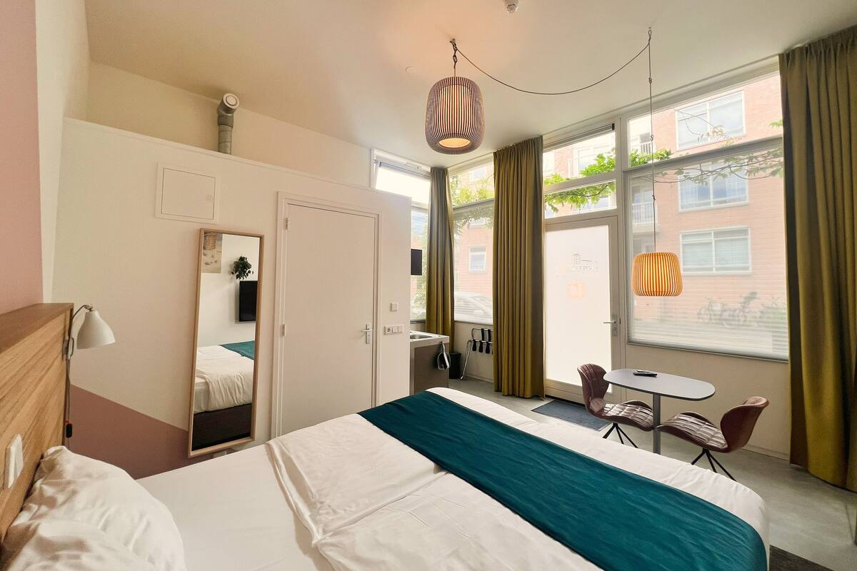 Quarto de hotel com cama, escrivaninha, cadeira, espelho e janela com cortinas.