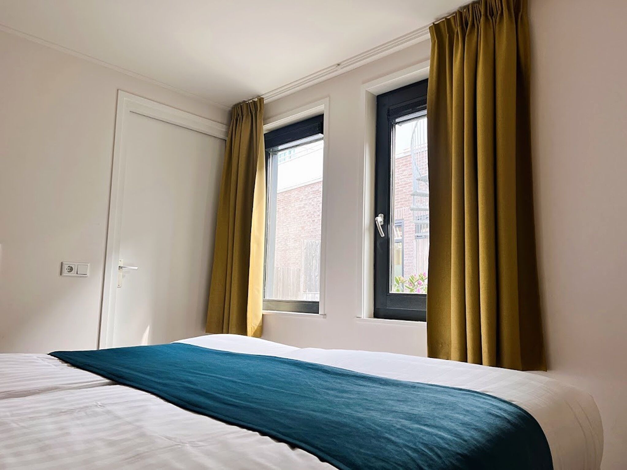 A hotel room with a bed, two windows with curtains, a door, and a view of the outside.