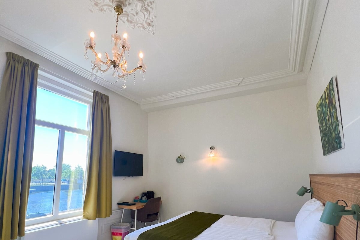 Quarto com cama, escrivaninha, cadeira, um banquinho pequeno, janela com cortinas e um lustre.