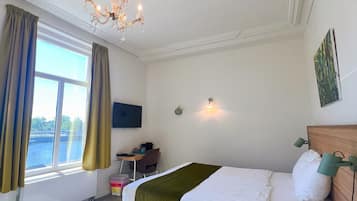 A bedroom with a bed, a desk, a chair, a small stool, a window with curtains, and a chandelier.