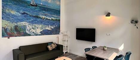 Una sala moderna con un cuadro grande de mar, un sofá verde oscuro, una mesita redonda y un comedor con sillas y mesa.
