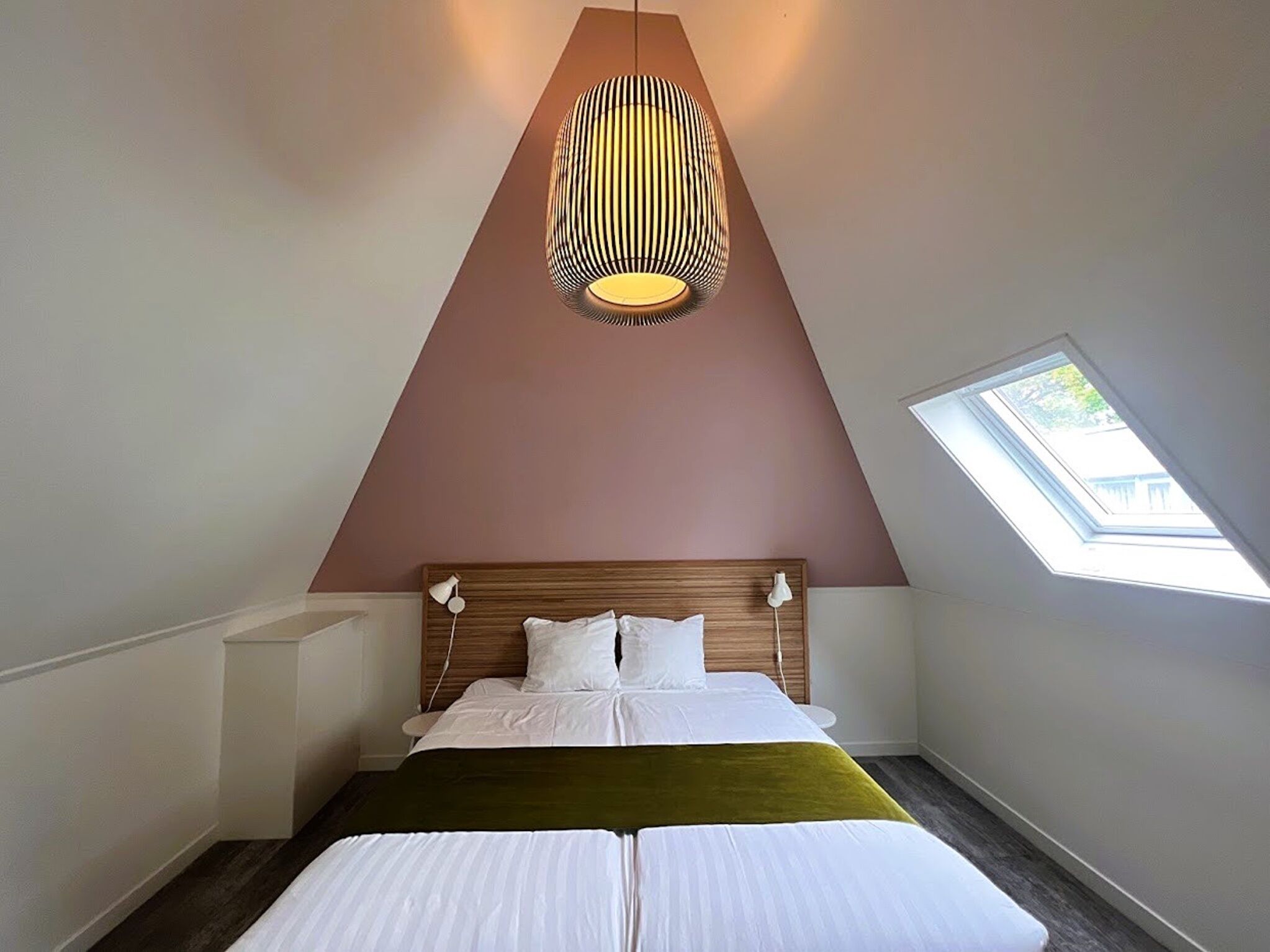 Un dormitorio con techo inclinado, tragaluz, cama con ropa blanca y lámpara colgante.