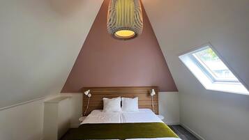 Un dormitorio con techo inclinado, tragaluz, cama con ropa blanca y lámpara colgante.