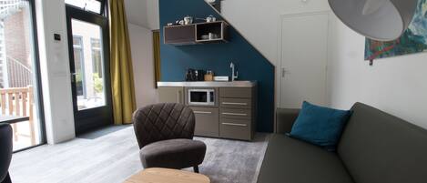 Een moderne woonkamer met een bank, een bijzettafeltje en een stoel. Er is een keukenhoek met een plank boven het aanrecht.