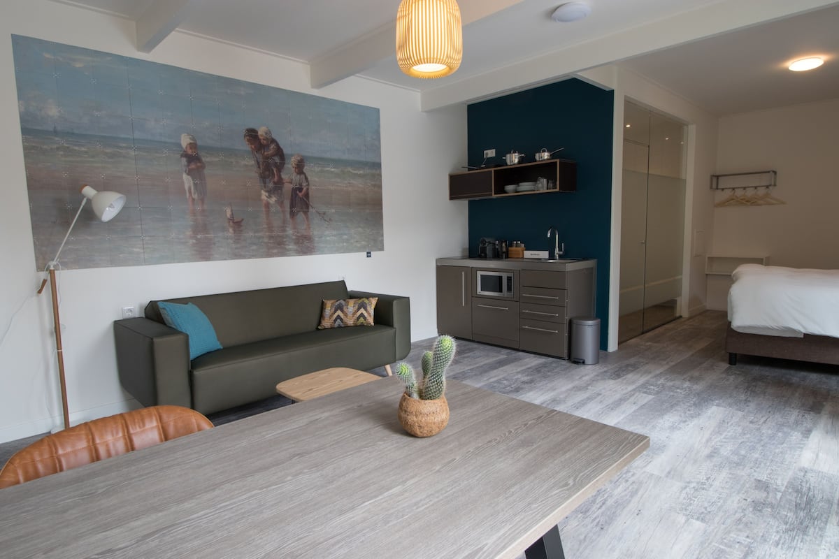 Uma sala de estar moderna com uma mesa de jantar de madeira, um sofá, uma pintura com uma cena de praia e uma área de cozinha com armários e um varal.