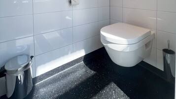 Un baño moderno con azulejos blancos, inodoro, un cubo de basura y lavamanos.