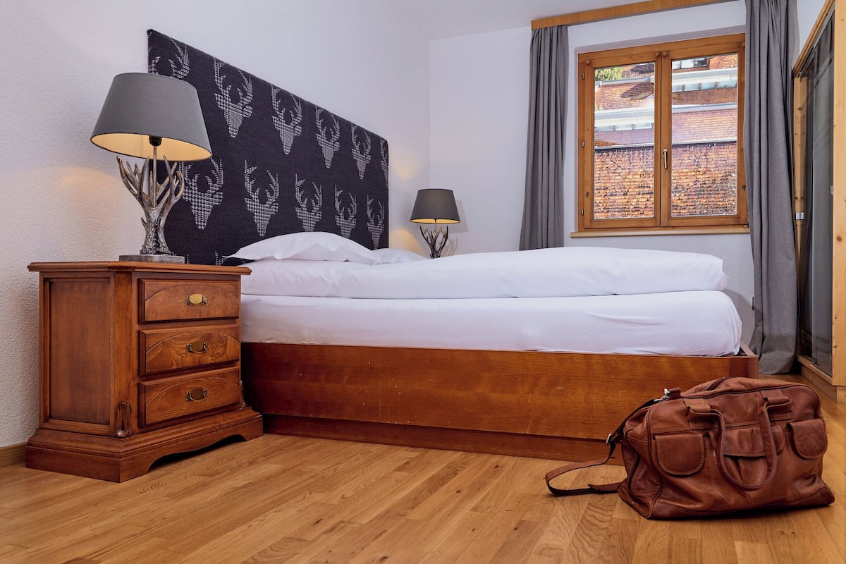 A bedroom with a wooden bed, a nightstand with a lamp, a dresser, and a window with curtains.