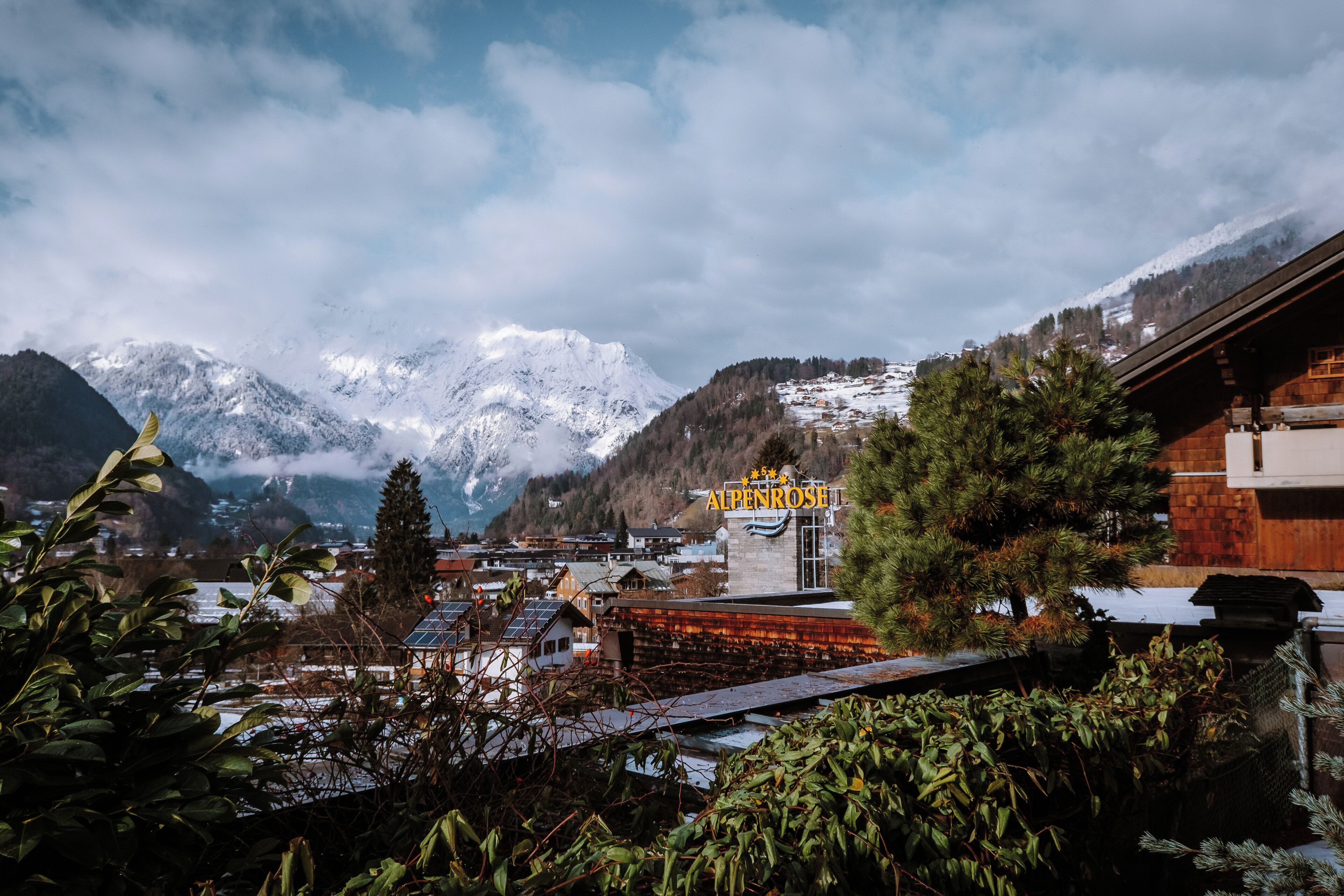 Un pueblo de montaña con picos nevados, un edificio de madera con el letrero 'ALPENROSE' y vistas del pueblo abajo.