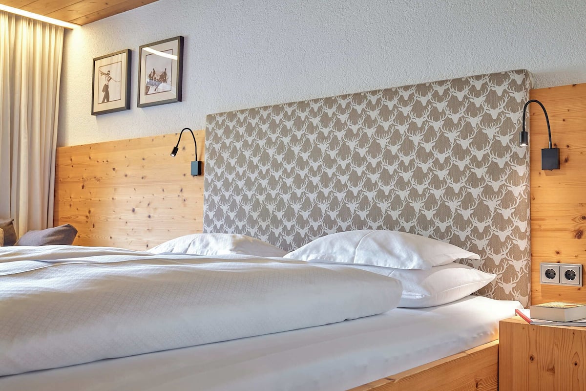 A neatly made bed with white linens, a wooden headboard with a patterned design, two wall-mounted lamps, and a nightstand with a power outlet.