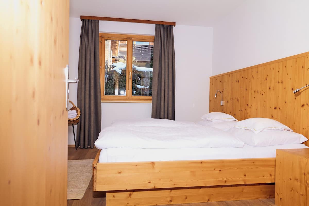 A wooden bed with white linens in a room with a window, a wooden headboard, and a wicker chair.