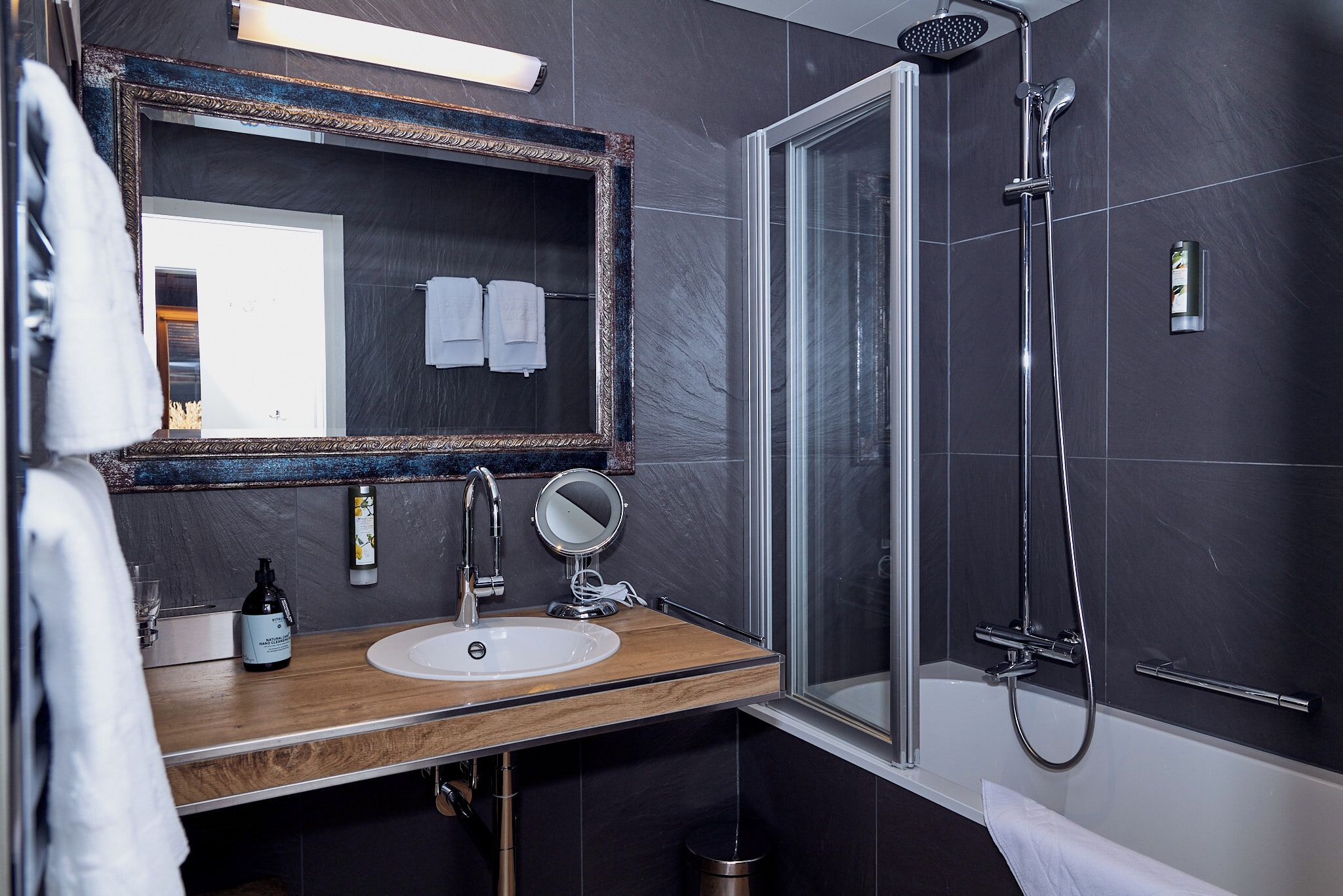 Une salle de bain moderne avec un meuble-lavabo en bois, un grand miroir, une douche à parois de verre et des murs recouverts de carreaux foncés.