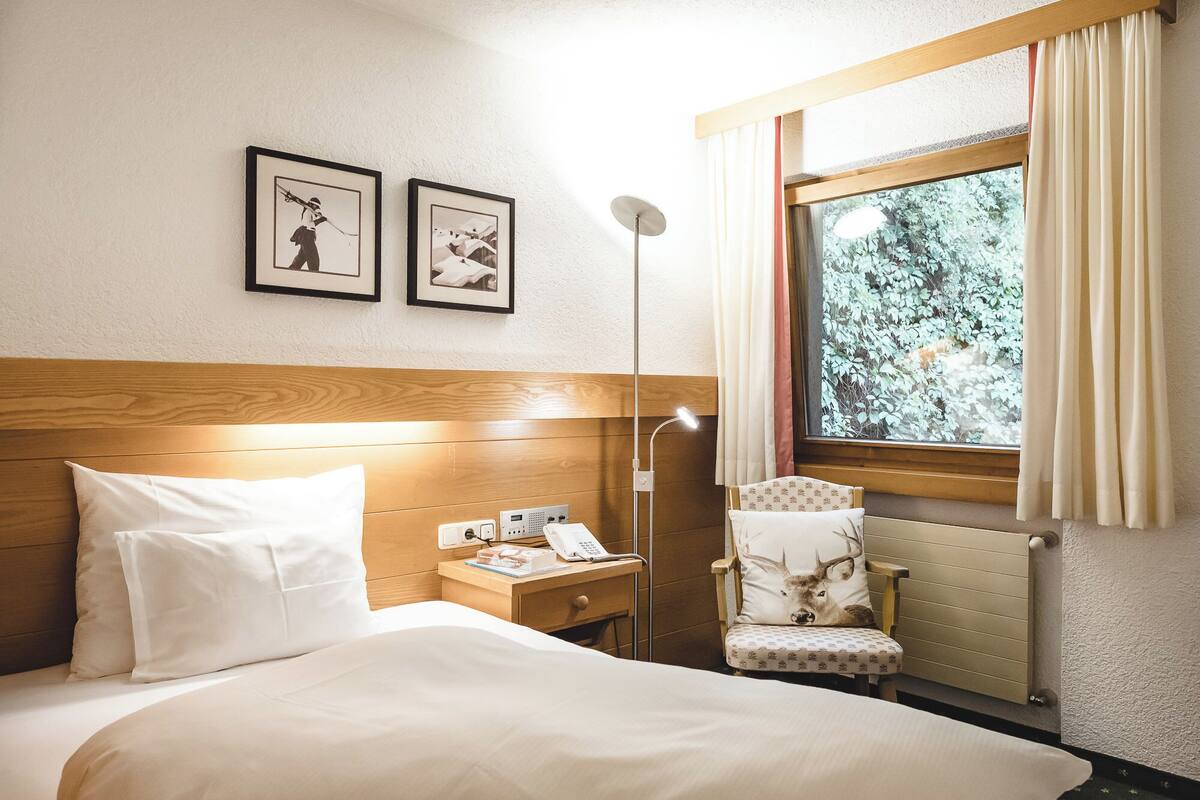 A hotel room with a bed, a nightstand with a phone, a lamp, a chair with a deer pillow, and a window with curtains.