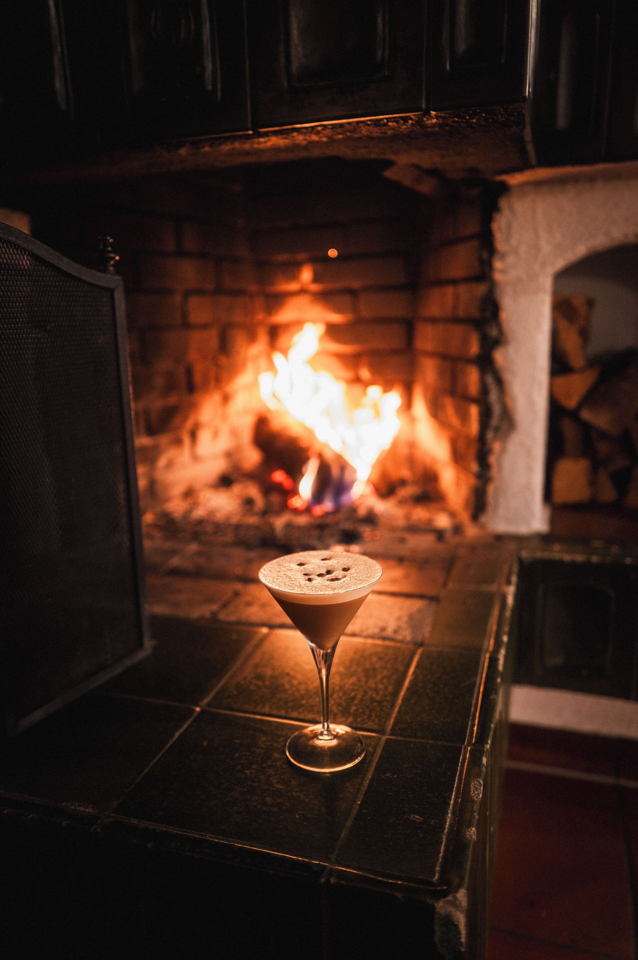 A martini glass on a table in front of a fireplace with a visible fire.