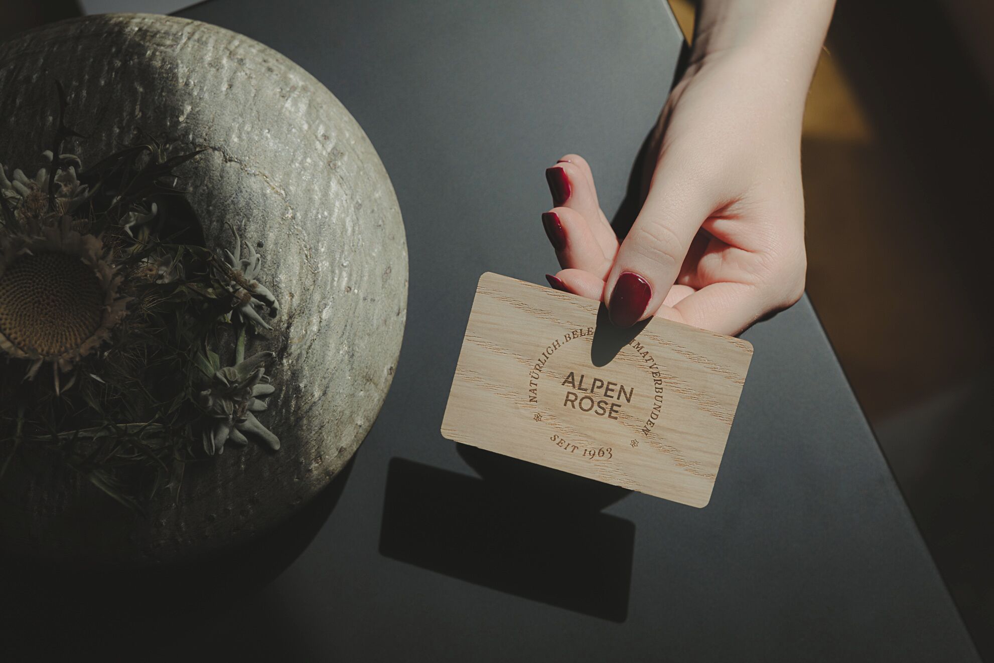 A hand holding a wooden card with 'ALPEN ROSE' and 'NATURLICH.BEILEIT 1963' text.