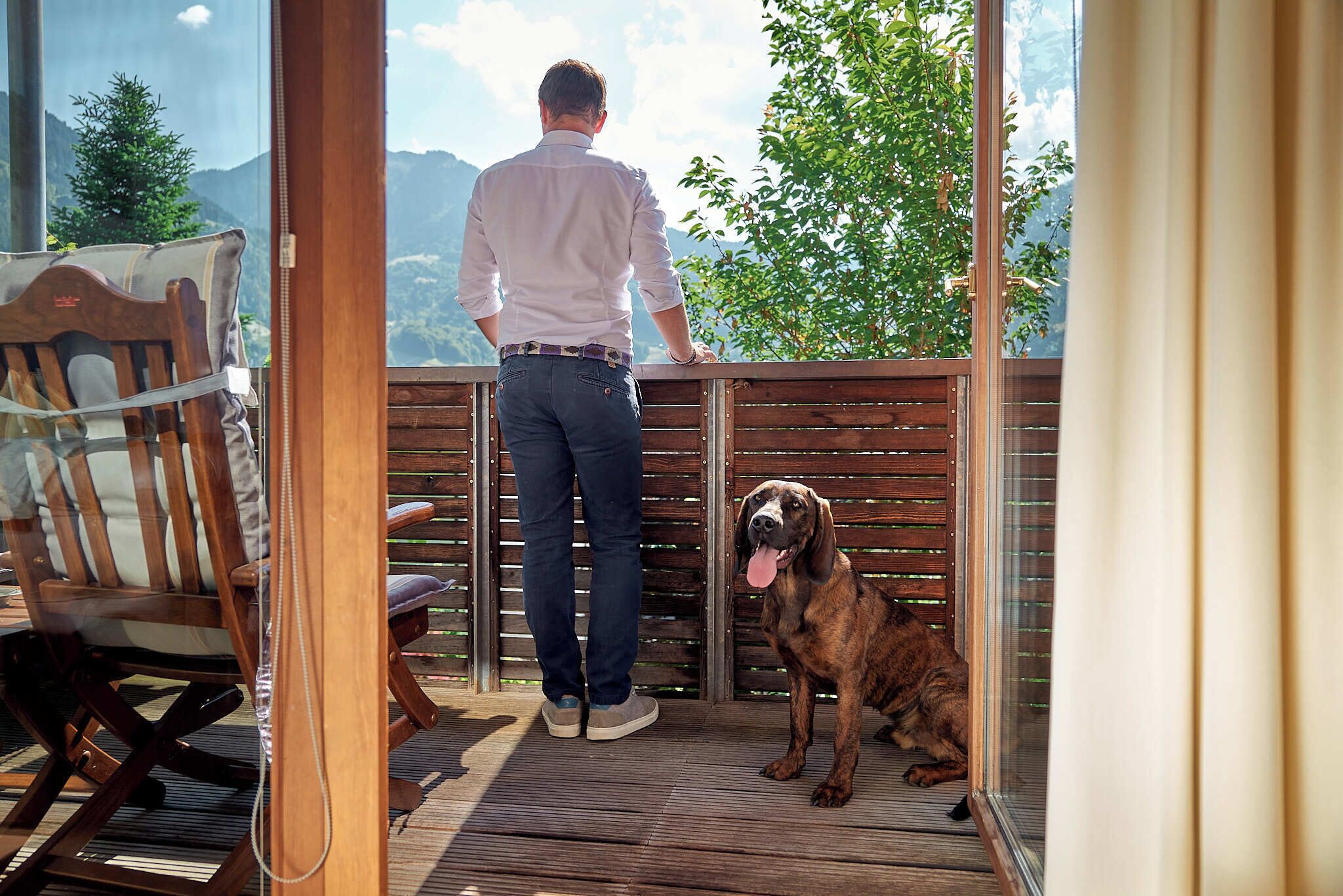 A man standing on a balcony overlooking a mountainous landscape, with a dog sitting beside him.