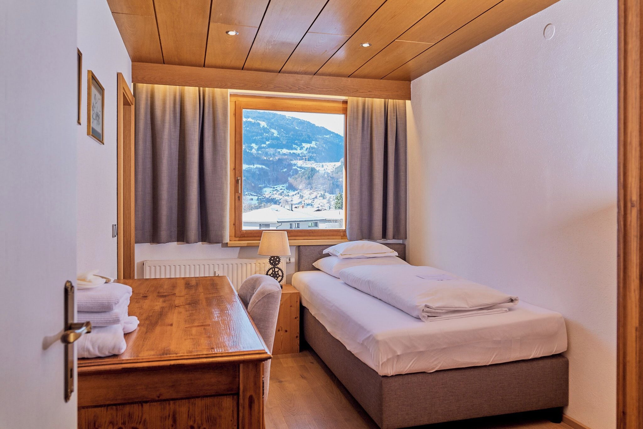 A small, cozy room with a wooden ceiling, a bed, a desk, and a window with curtains.