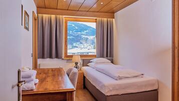 A small, cozy room with a wooden ceiling, a bed, a desk, and a window with curtains.