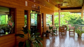 A wooden-floored patio with potted plants, a seating area with chairs, and a view of greenery outside.