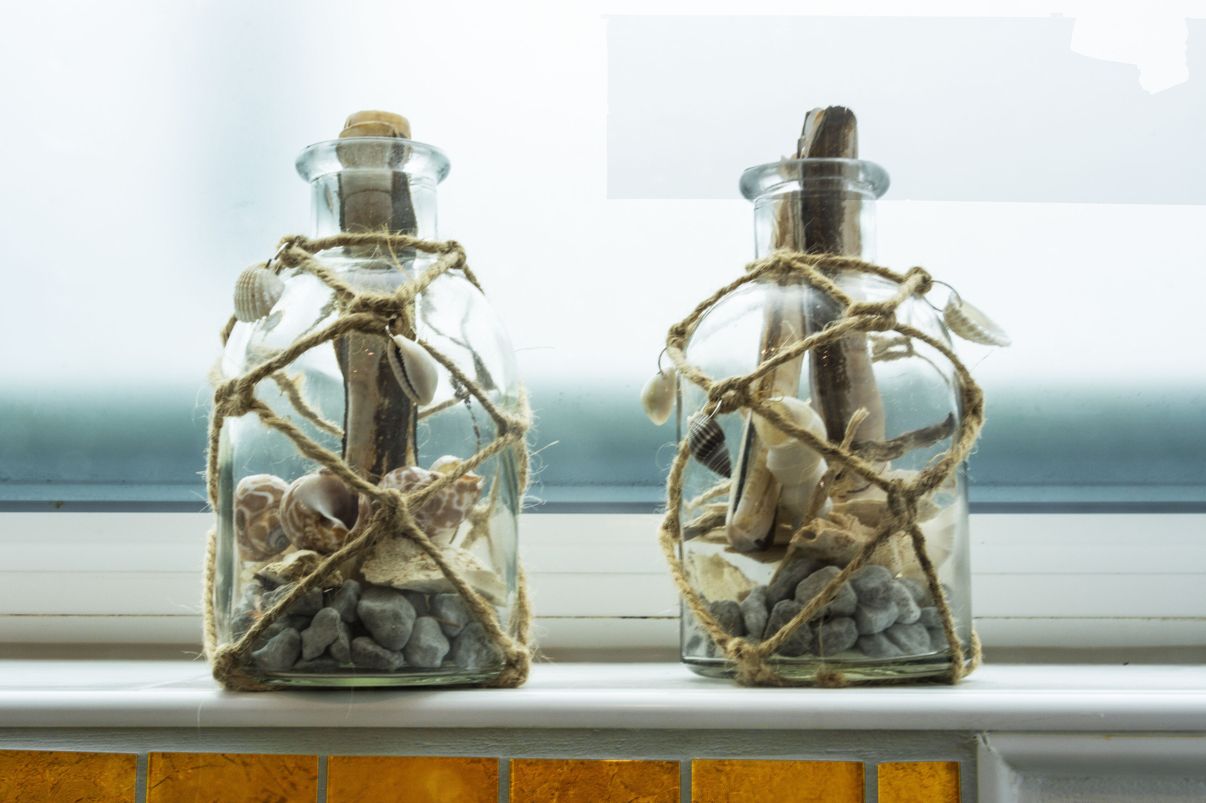 Deux bouteilles en verre, décorées de ficelle et de coquillages, sont posées sur un rebord de fenêtre.