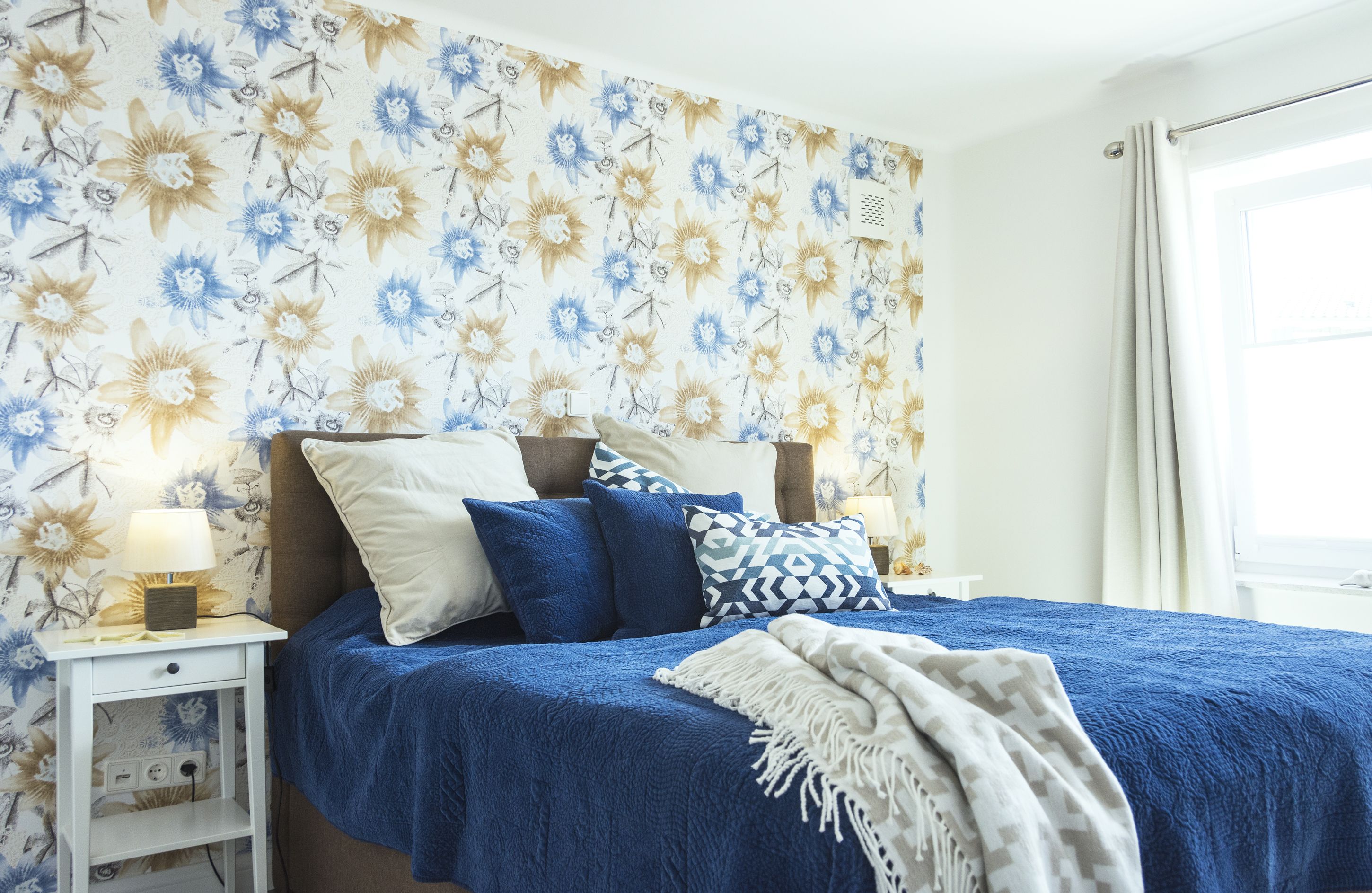 A bedroom with a floral wallpaper, a blue bedspread, and a bedside table with a lamp.
