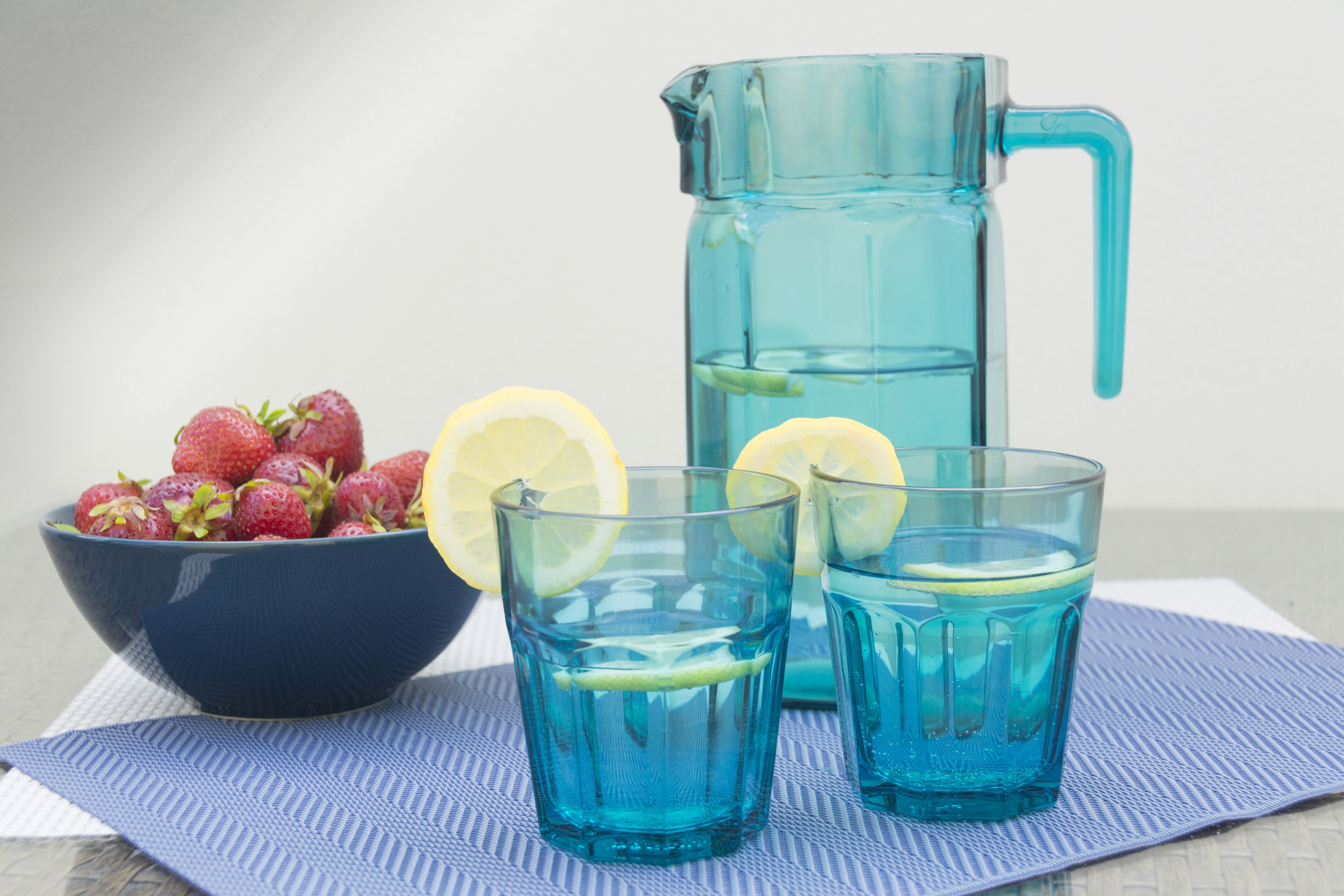 A pitcher, two glasses, a bowl of strawberries, and a lemon slice on a blue mat.