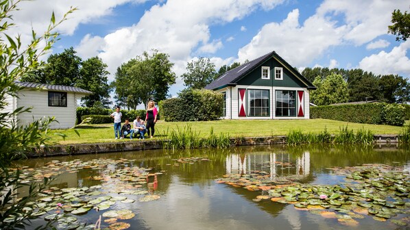 A house with a pond and lily pads, surrounded by greenery and a clear sky.
