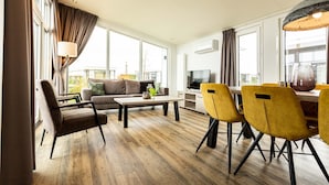 Een moderne woonkamer met een bank, salontafel en gele stoelen. Er is een televisie en een eettafel met stoelen.