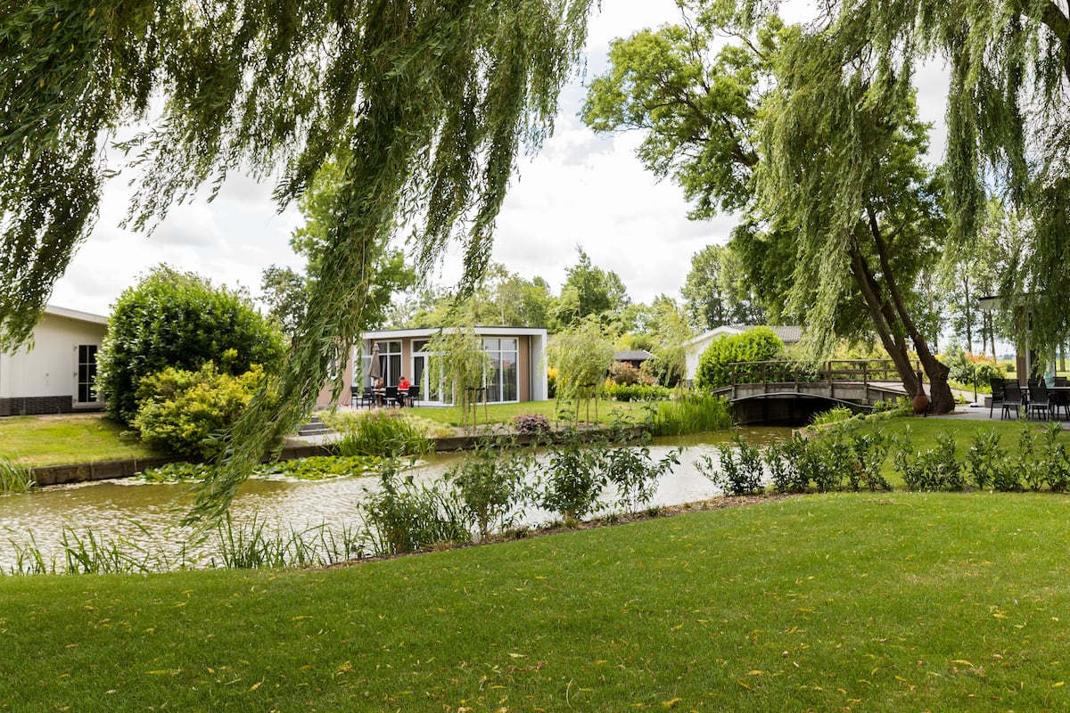 Un jardín tranquilo con un estanque, un puente y un edificio moderno con mesas al aire libre.
