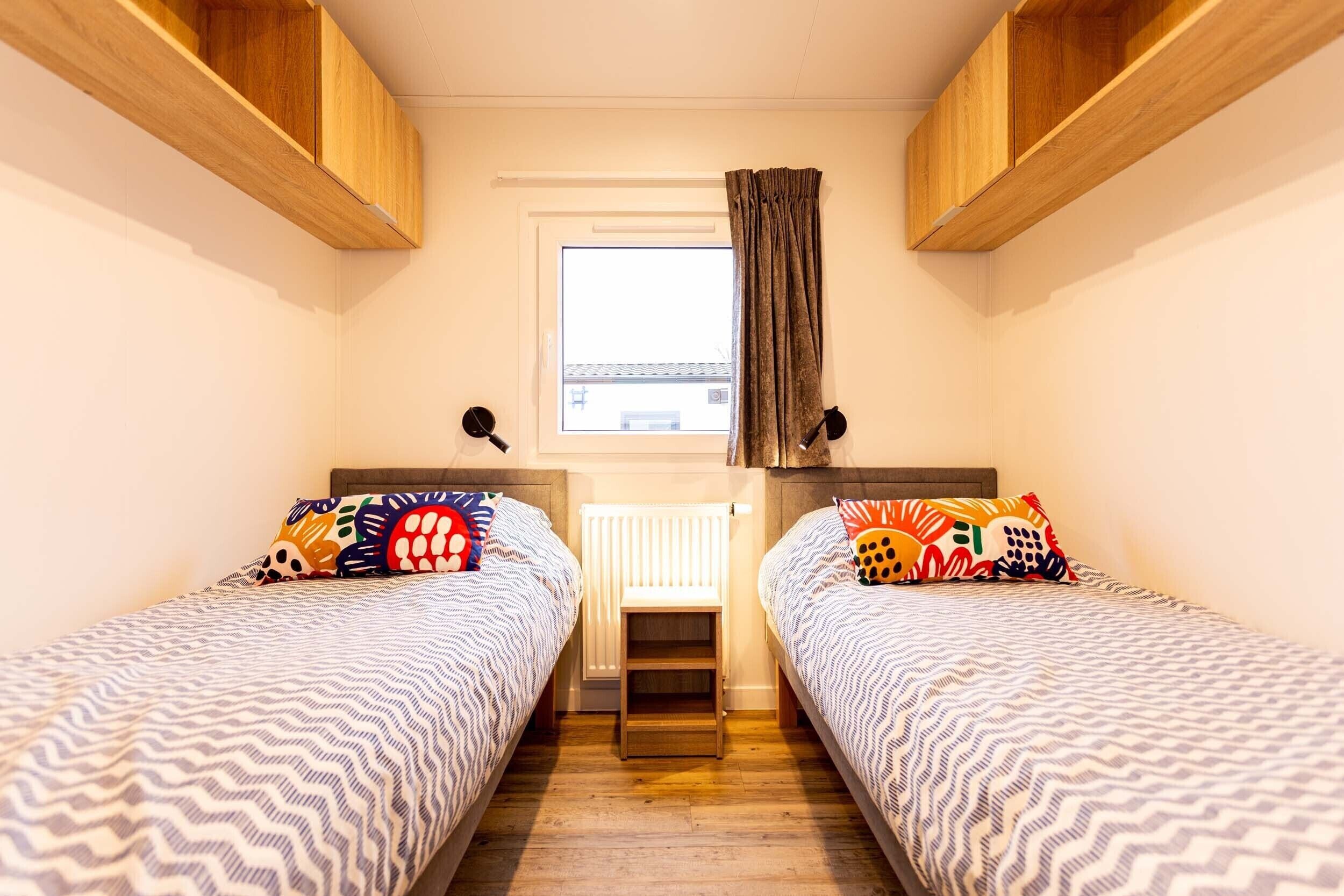 A small, cozy room with two single beds, a window with curtains, and wooden shelves above the beds.