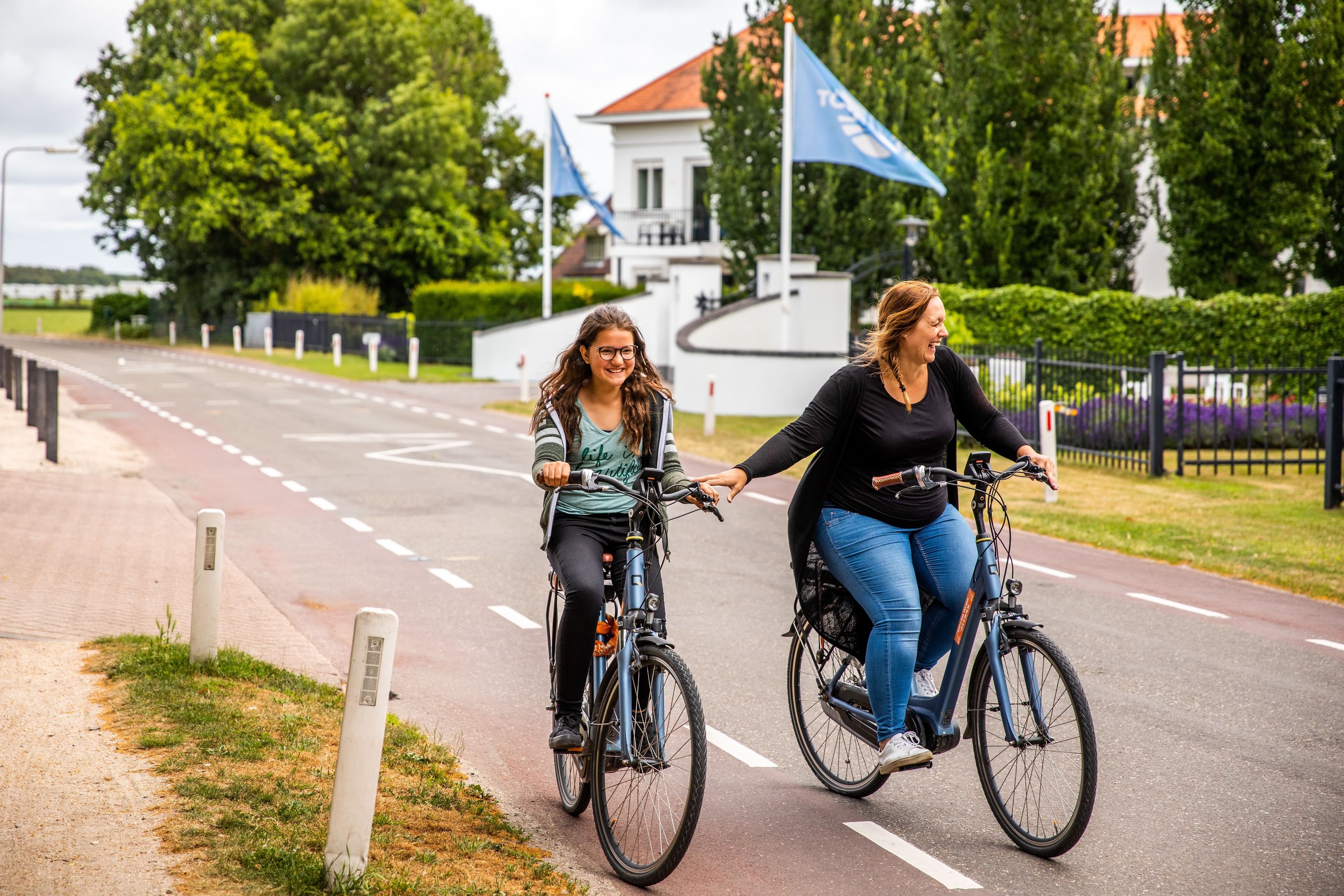 Two people riding bicycles on a paved path with a building and flag in the background.