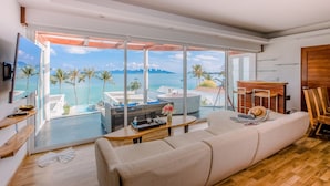 A modern living room with a large window overlooking a pool and ocean. There is a sofa, a coffee table, and a TV mounted on the wall. - The Privilege Hotel Ezra Beach Club (Koh Samui)