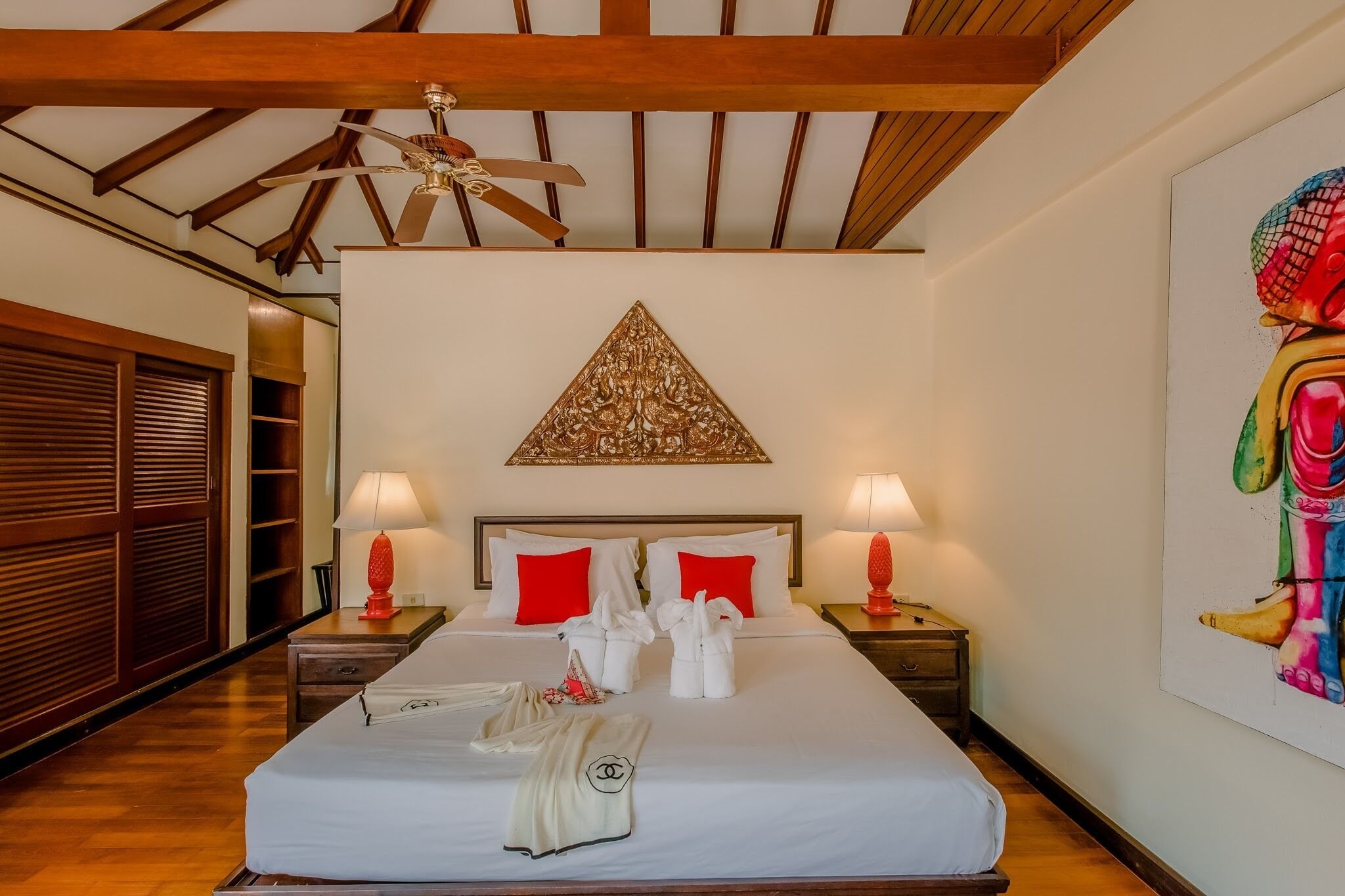 A hotel room with a large bed, wooden ceiling beams, a ceiling fan, two bedside lamps, a red and white pillow set, a decorative wall piece, and a large painting on the wall.