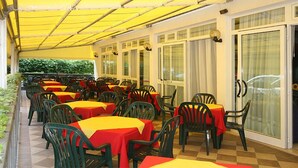 Un espace de restauration en plein air, avec des tables et des chaises, abrité par un auvent jaune, et doté de grandes fenêtres sur un côté.