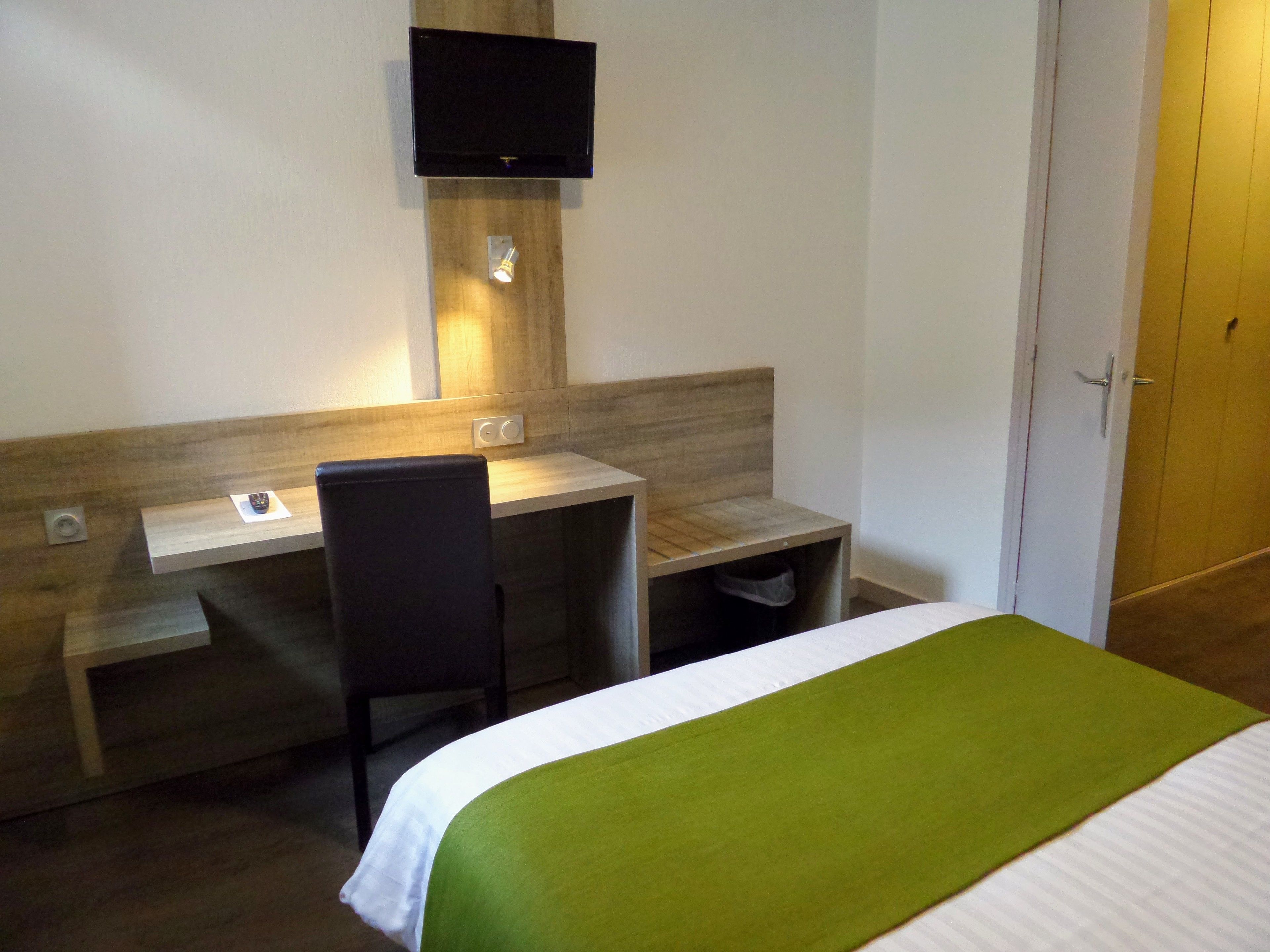 A hotel room with a bed, a desk, a chair, a television mounted on the wall, and a door.
