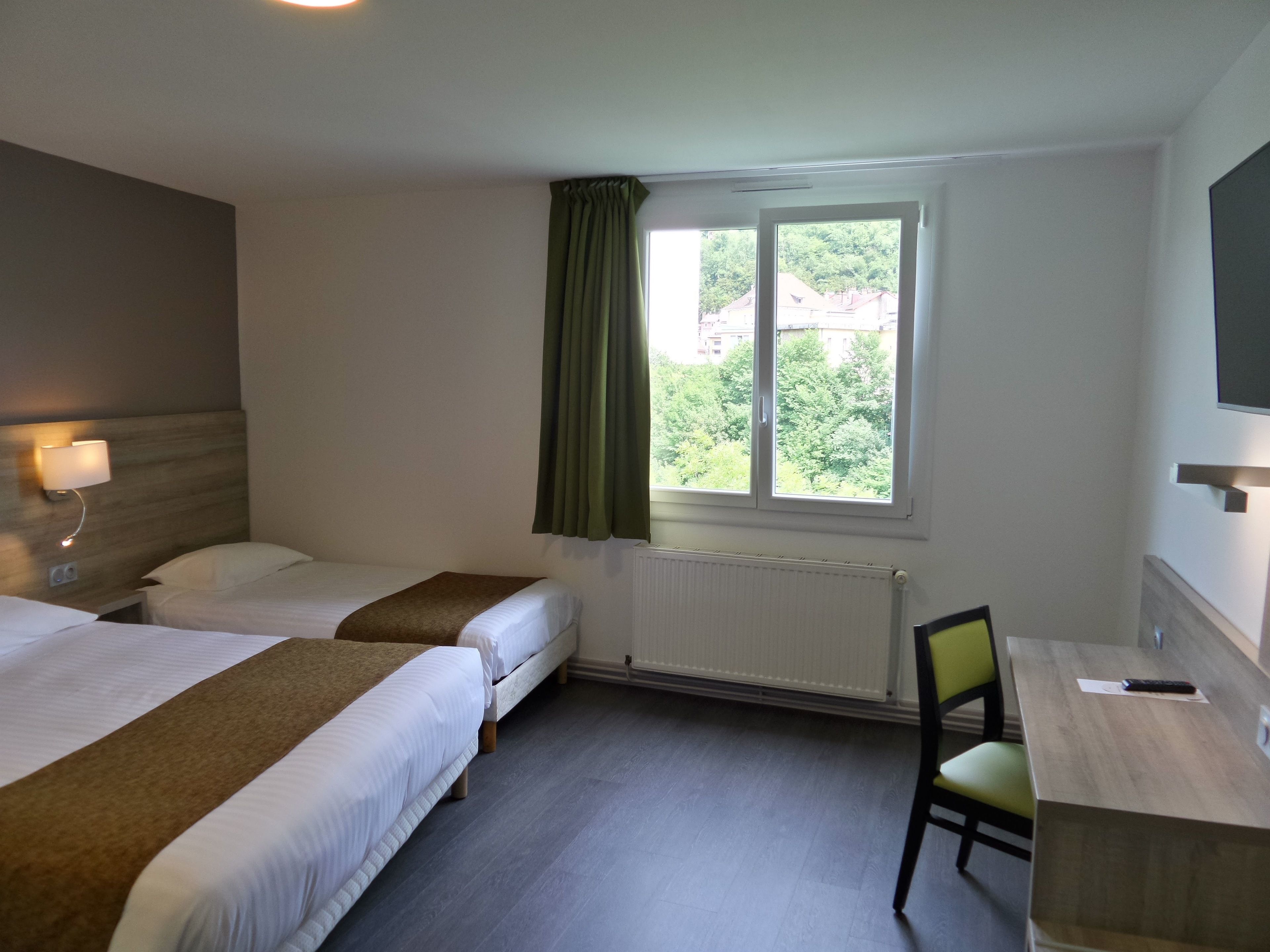 Ein Hotelzimmer mit zwei Betten, einem Schreibtisch, einem Stuhl, einem Fenster mit Vorhängen und einem an der Wand befestigten Fernseher.