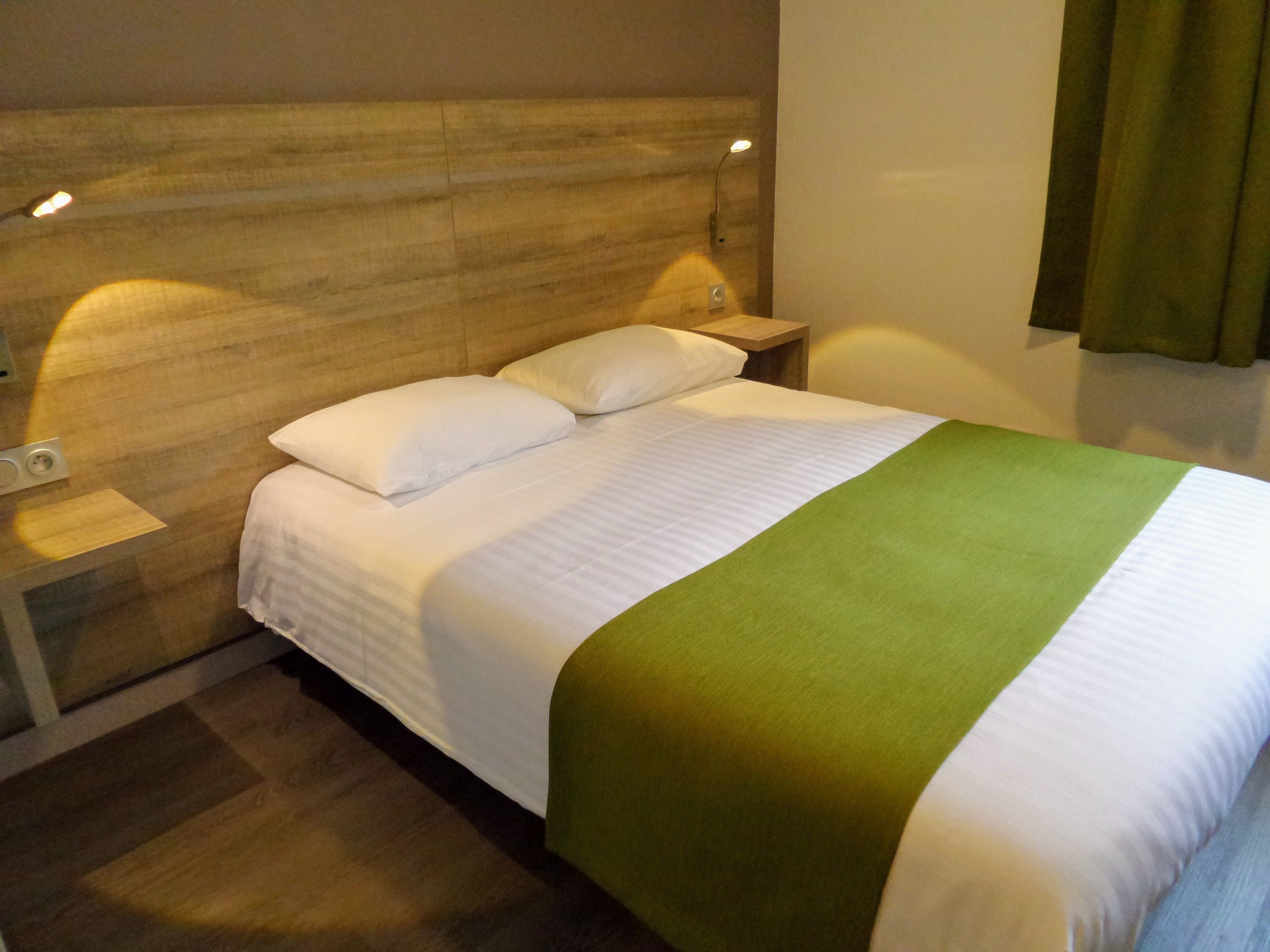 Un lit bien fait, avec du linge de lit blanc et une touche de vert, une tête de lit en bois, une lampe de chevet et une table de chevet.
