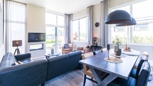 Una sala de estar moderna con sofá, mesa de comedor y sillas. Hay un televisor montado en la pared y una ventana con cortinas.