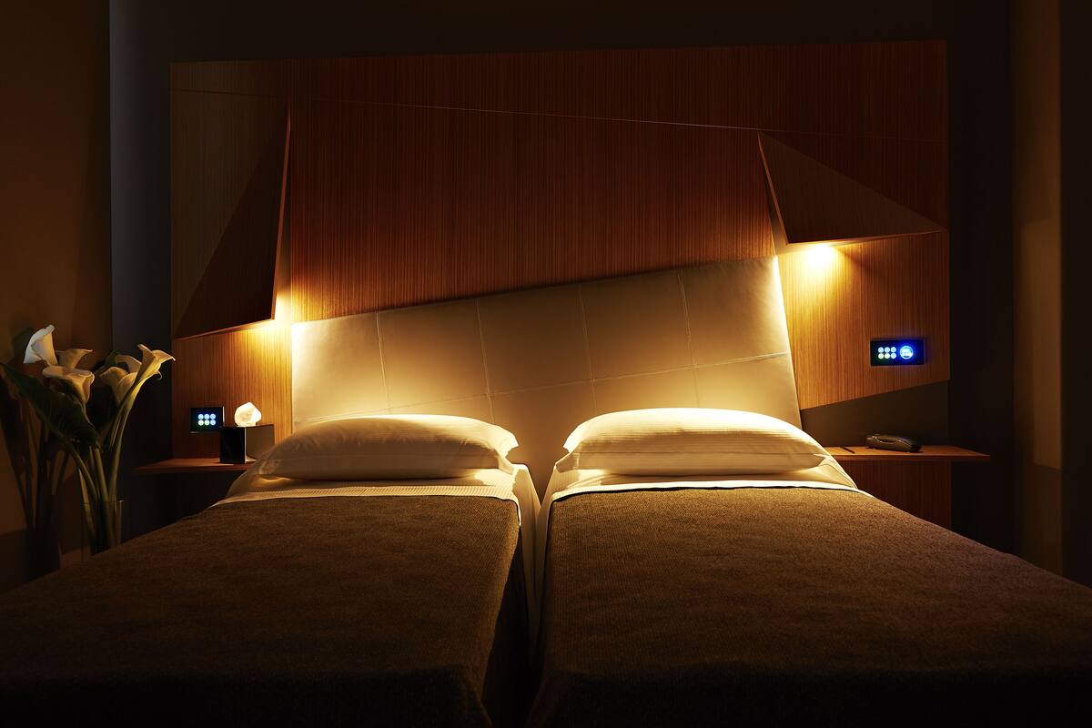 A hotel room with two beds, a nightstand with a lamp, and a wall-mounted control panel.