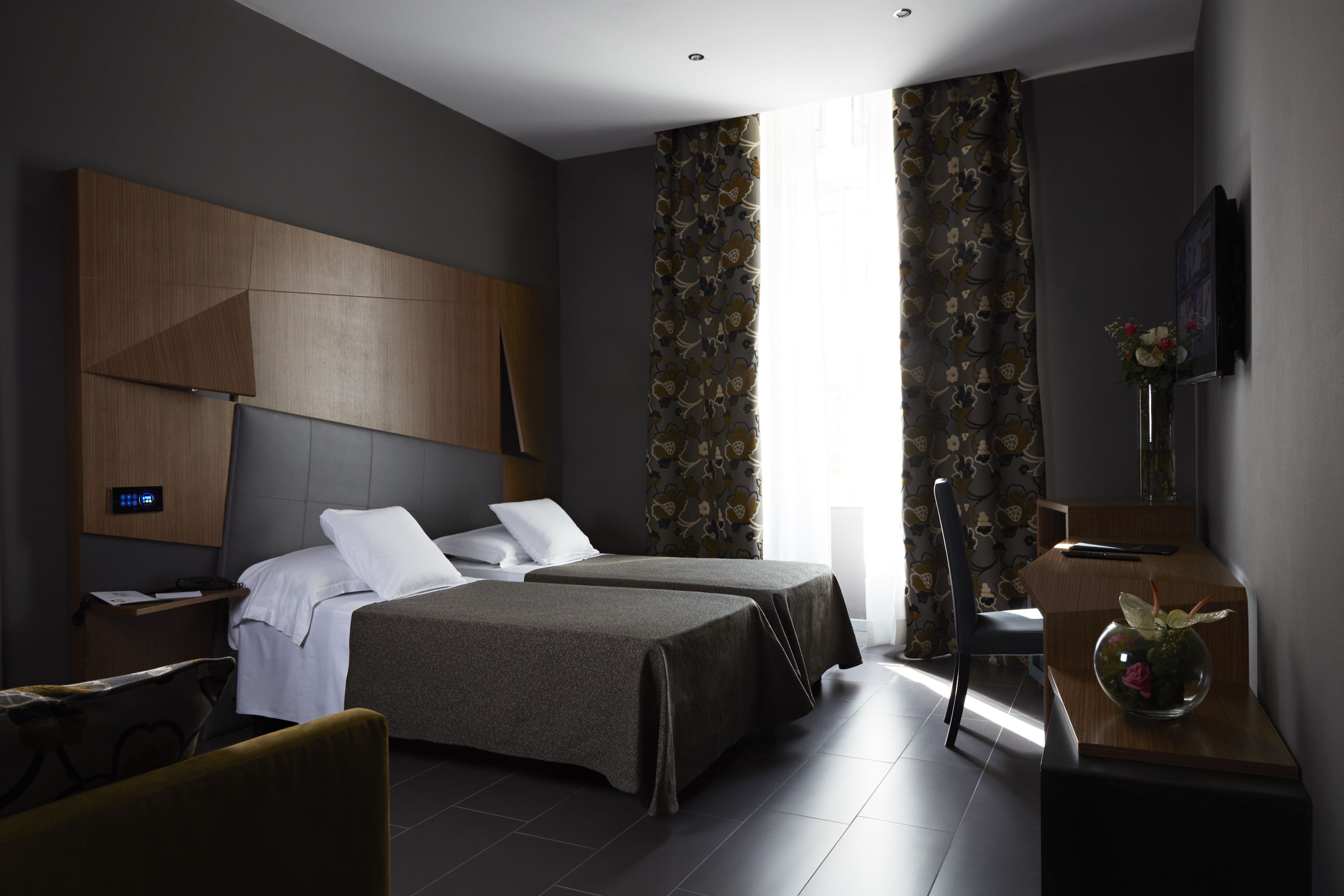 A modern hotel room with two beds, a wooden headboard, a desk with a vase, and a large window with curtains.