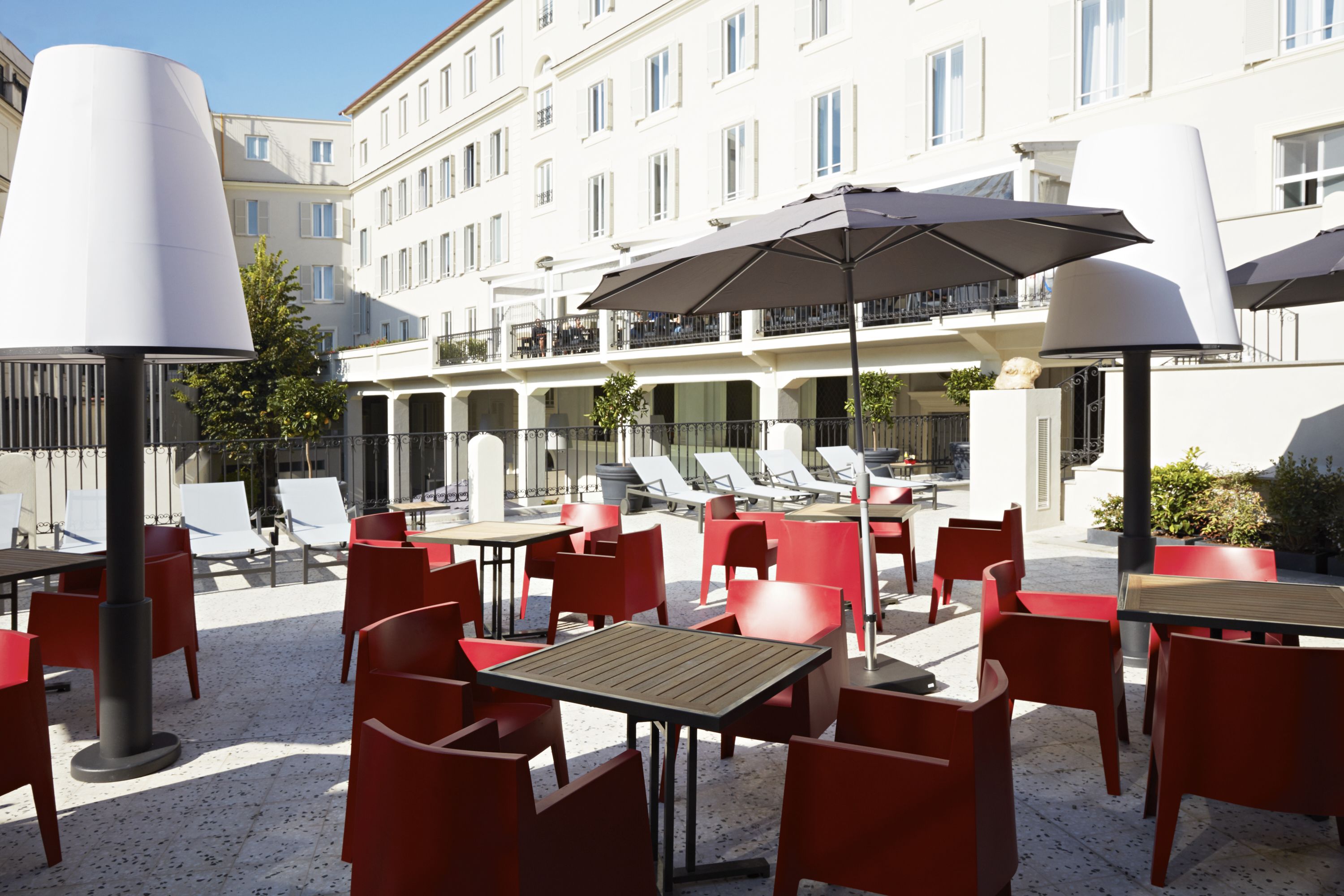 An outdoor cafe area with red chairs and tables, umbrellas, and a view of a building with balconies.