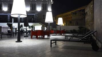An outdoor seating area with tables and chairs, illuminated by large lamps, in front of a building with a staircase and a stone wall.